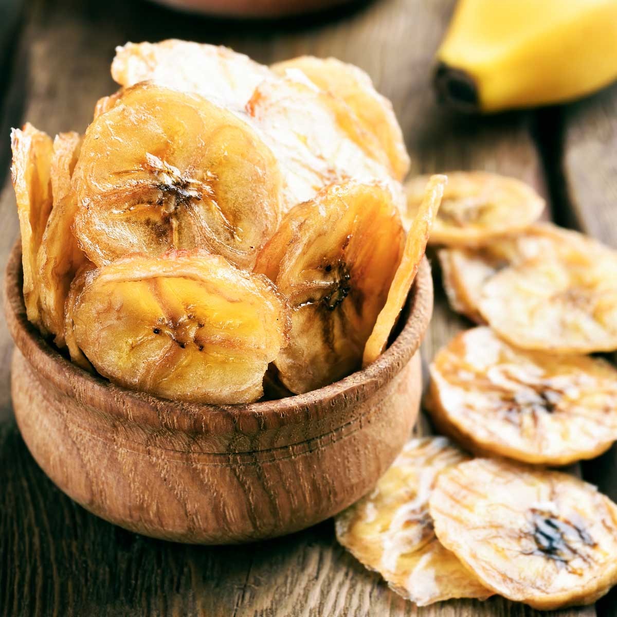 Desipan crispy dehydrated banana chips in a rustic wooden bowl on a dark wooden surface with a fresh banana nearby.