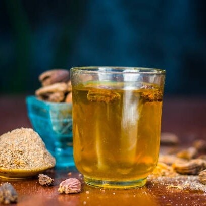 Desipan Herbal Infusion Beverage With Spices, golden liquid in a glass with black cardamom pods and powder.