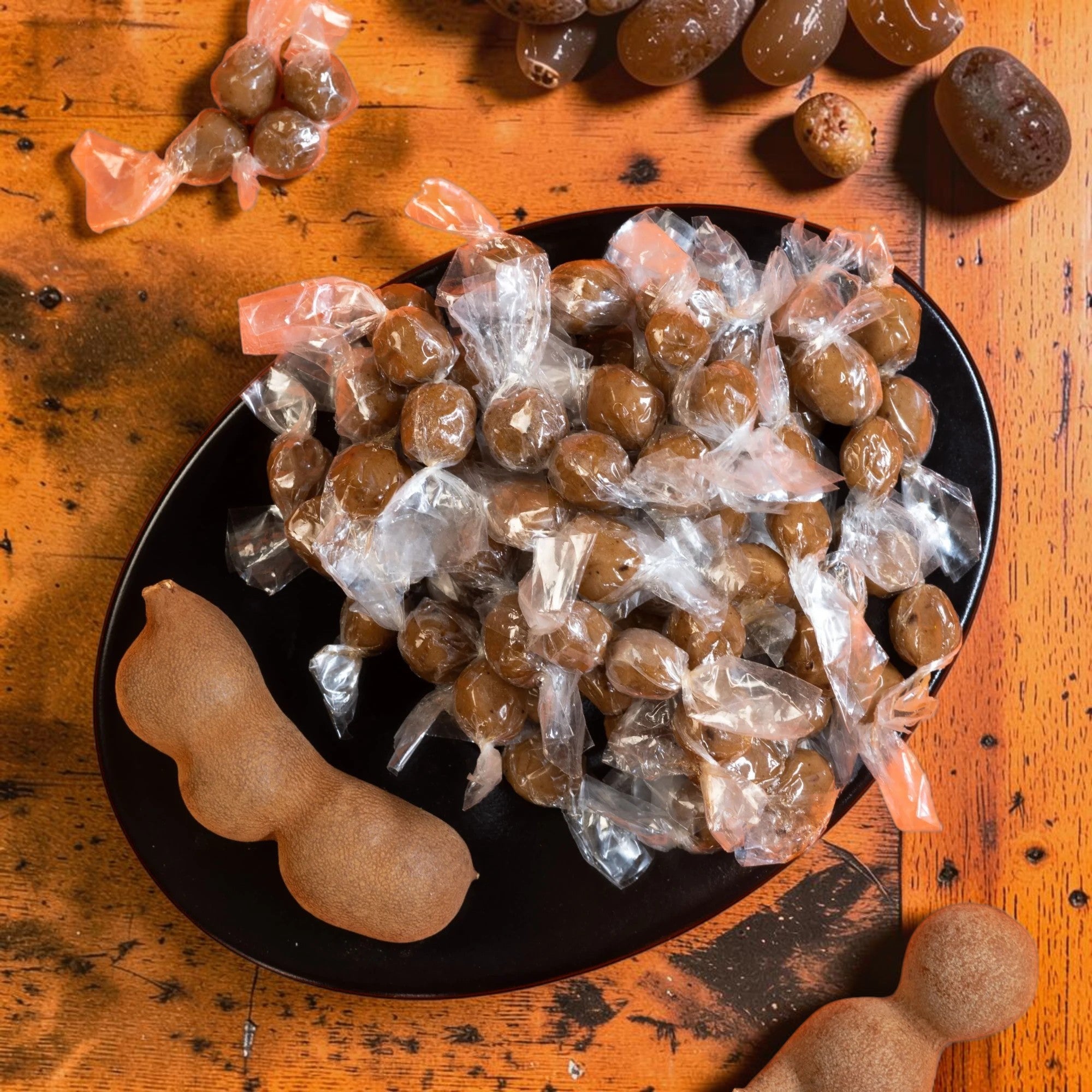 Desipan Tamarind candies, individually wrapped, on a black plate with tamarind pods. Sweet and tangy tropical flavor.